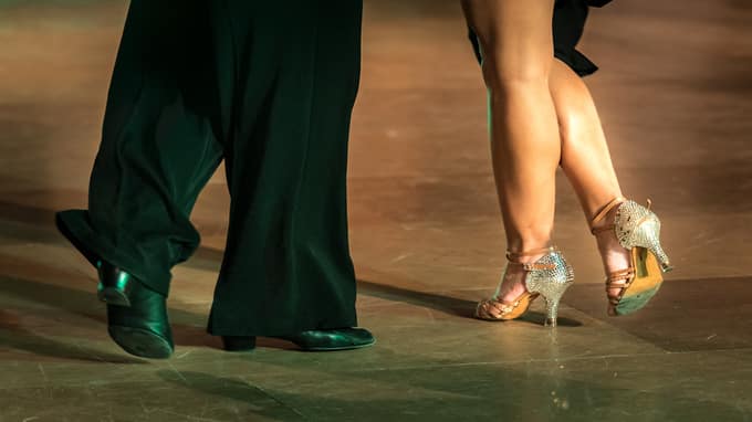 Pieds de danseurs exécutant un pas sur un parquet. Un partenaire porte des chaussures noires, l'autre des talons hauts scintillants.