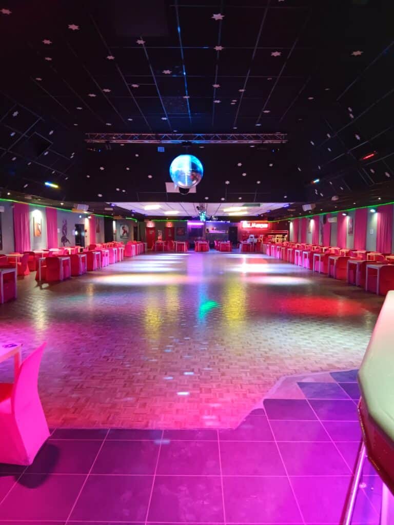Salle de danse festive avec plafond étoilé, boule à facettes, rideaux roses, tables rouges et lumières colorées sur parquet bois. Texte : LE MARCQ.