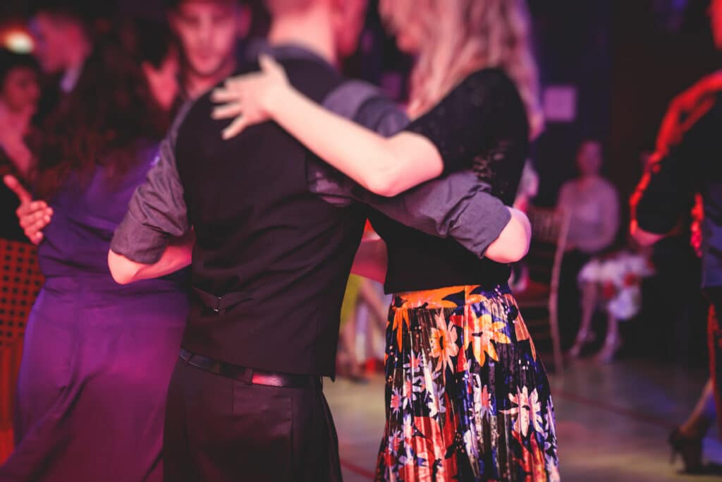Couples dansant élégamment lors d'une soirée, éclairés par des lumières colorées. Une danseuse porte une jupe plissée florale.