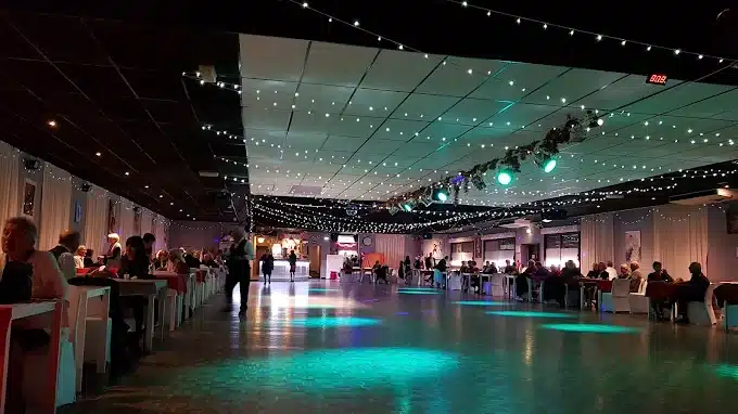 Salle de bal illuminée pour une soirée dansante. Des guirlandes lumineuses, des spots verts et un public assis autour d'une grande piste. Heure: 8:02.