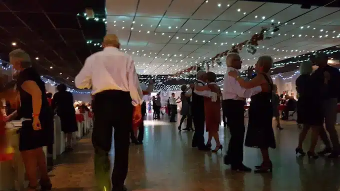 Des couples dansent dans une grande salle de bal éclairée par des lumières scintillantes, avec des tables en arrière-plan.