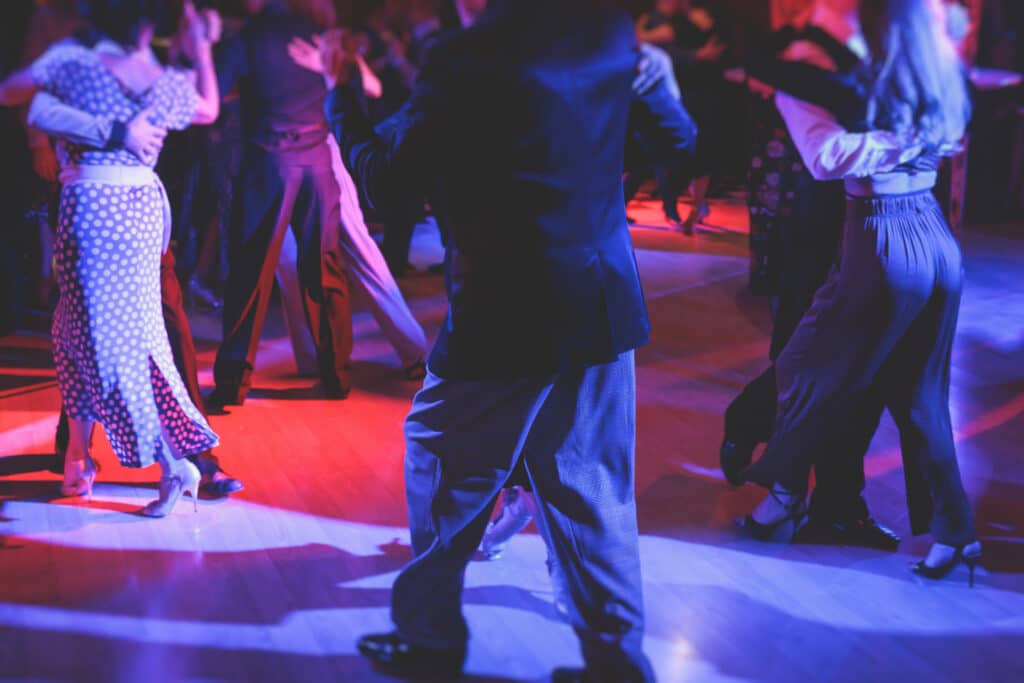 Couples élégants dansant le tango sur une piste en bois sous des lumières rouges et bleues. Robe à pois, costumes et talons hauts.